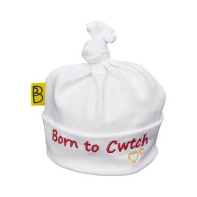 Handmade white Welsh organic cotton baby hat embroidered with words Born to Cwtch in red and two yellow embroidered hearts