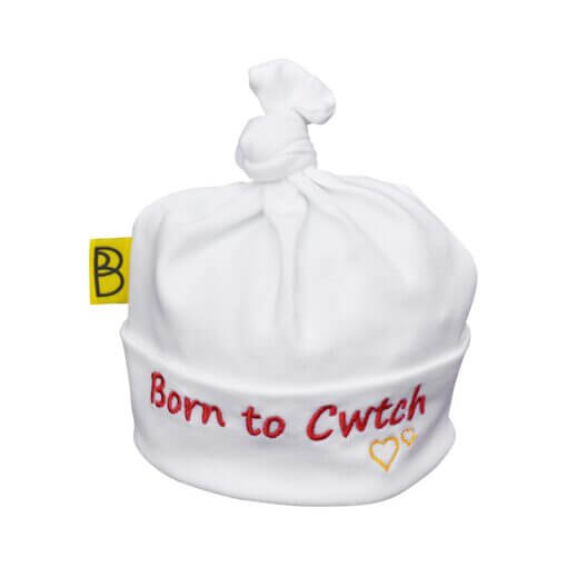 Handmade white Welsh organic cotton baby hat embroidered with words Born to Cwtch in red and two yellow embroidered hearts